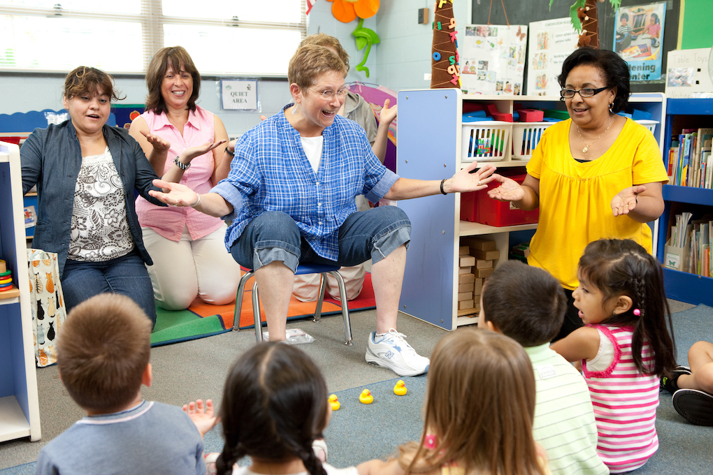 a teacher sitting in a small chair in the middle of a classroom is teaching a learning game to other educators, children and a teaching artist