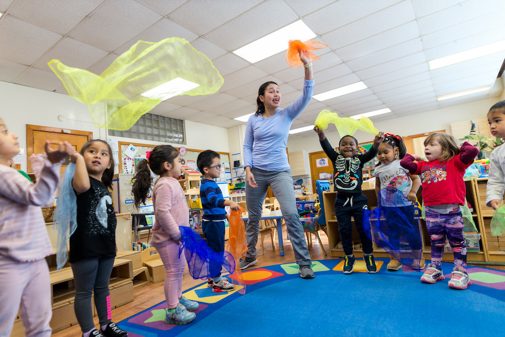 a wolf trap teaching artist is standing in a circle with young children in the middle of a classroom playing a learning game with colorful handkerchiefs