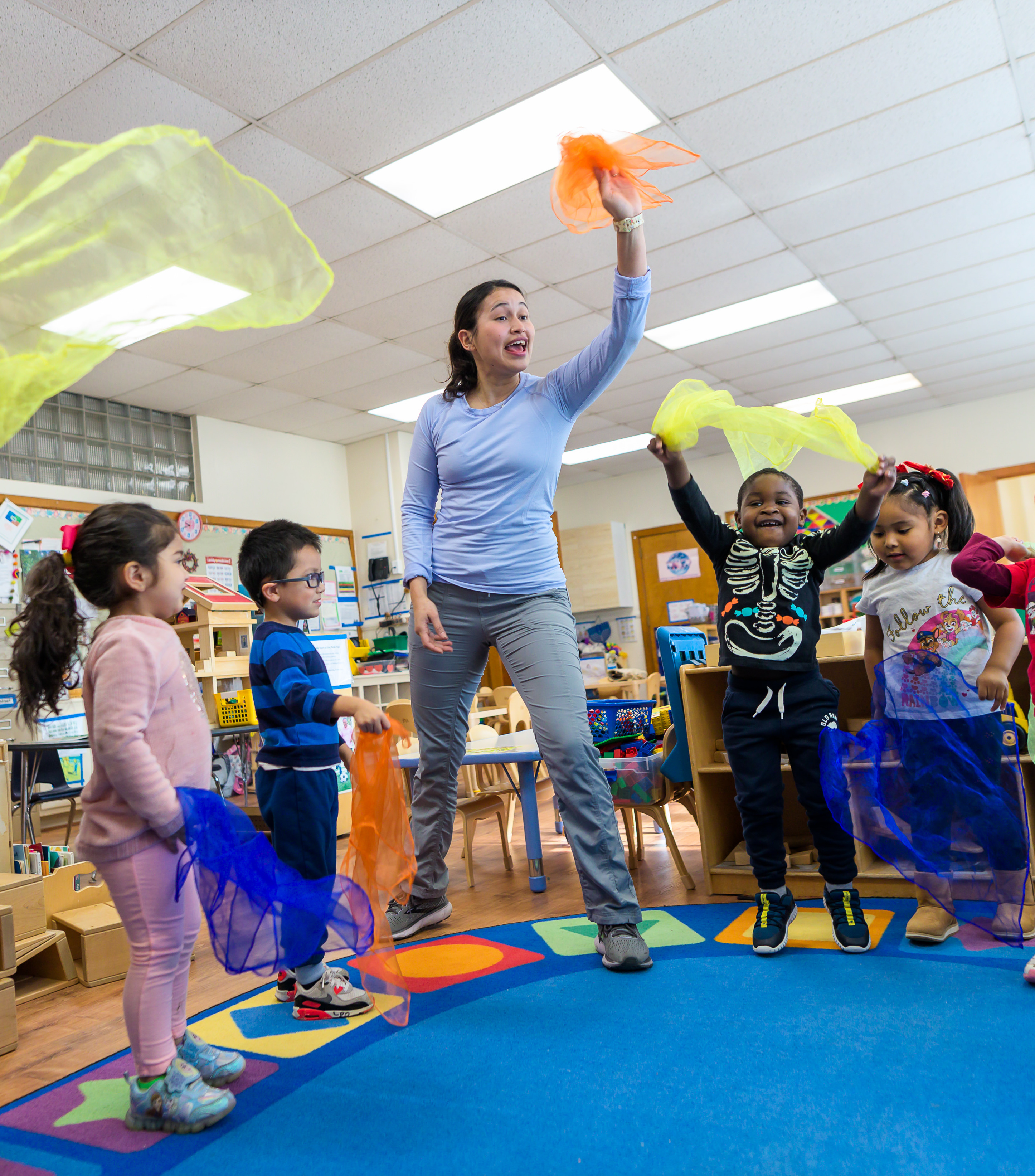 a wolf trap teaching artist is standing in a circle with young children in the middle of a classroom playing a learning game with colorful handkerchiefs