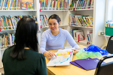 a wolf trap teaching artist and an educator are sitting at a library table while the wolf trap teaching artist shares classroom ideas from a colorful book