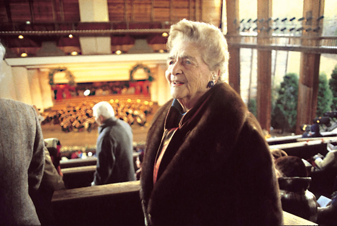 Catherine Filene Shouse a fur coat standing in a balcony area overlooking a holiday concert stage decorated with wreaths.