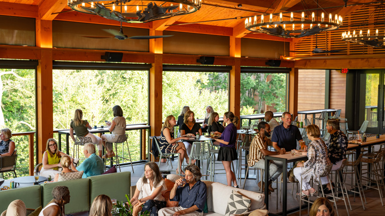 A wide view of Meadow Lounge, filled with chatting members. There are high and low tables, sofas, and chandeliers.