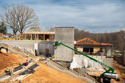 Large construction site with workers, a green boom lift, and multiple unfinished buildings surrounded by dirt and temporary barriers.