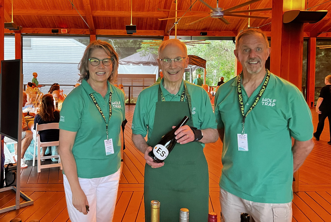 Three Wolf Trap volunteers in green shirts standing at a wooden pavilion, one holding a wine bottle with other bottles on the table.