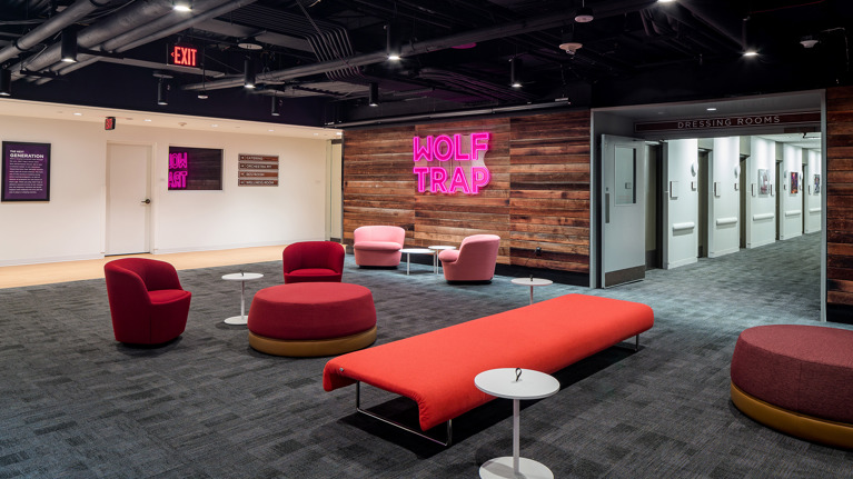 Reception area with modern seating, wood-paneled walls, and neon “Wolf Trap” sign near dressing room doors.