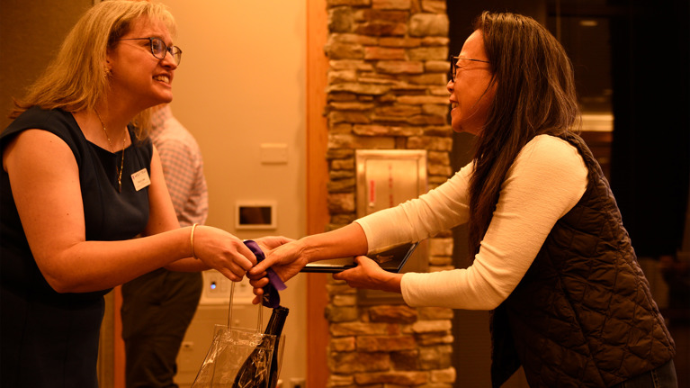 Wolf Trap staff member hands a gift bag and certificate to Lisa Hia near a stone accent wall, with “Wolf Trap” visible on the bag.