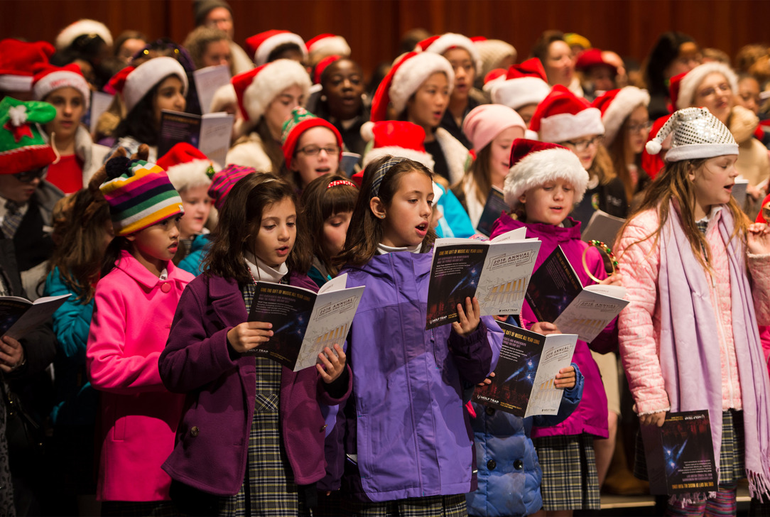 Group of children wearing colorful coats and Santa hats singing from songbooks on stage.