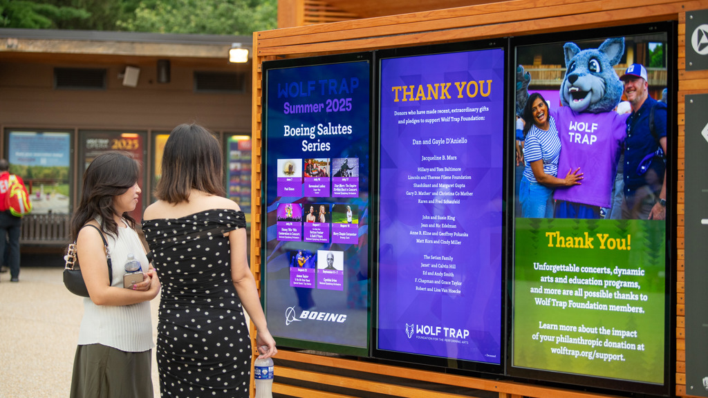 Two patrons on the Filene Center Plaza look at a Boeing performance series poster displayed on a screen.