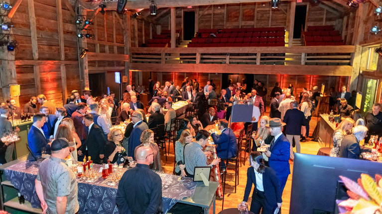 Large crowd mingling inside a rustic wooden venue with tables and bars.