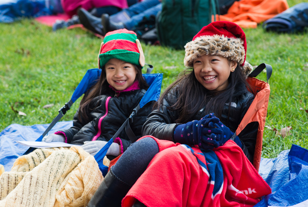 Two young girls sitting on lawn chairs with festive hats and blankets at an outdoor holiday event.