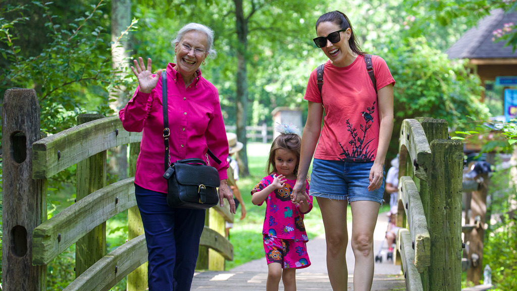 A grandmother, mother, and daughter walk on a wooden bridge in a green outdoor setting and wave to the camera.