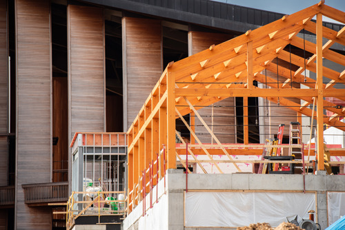 Close-up of a wooden frame structure under construction next to a large building with vertical wooden panels.
