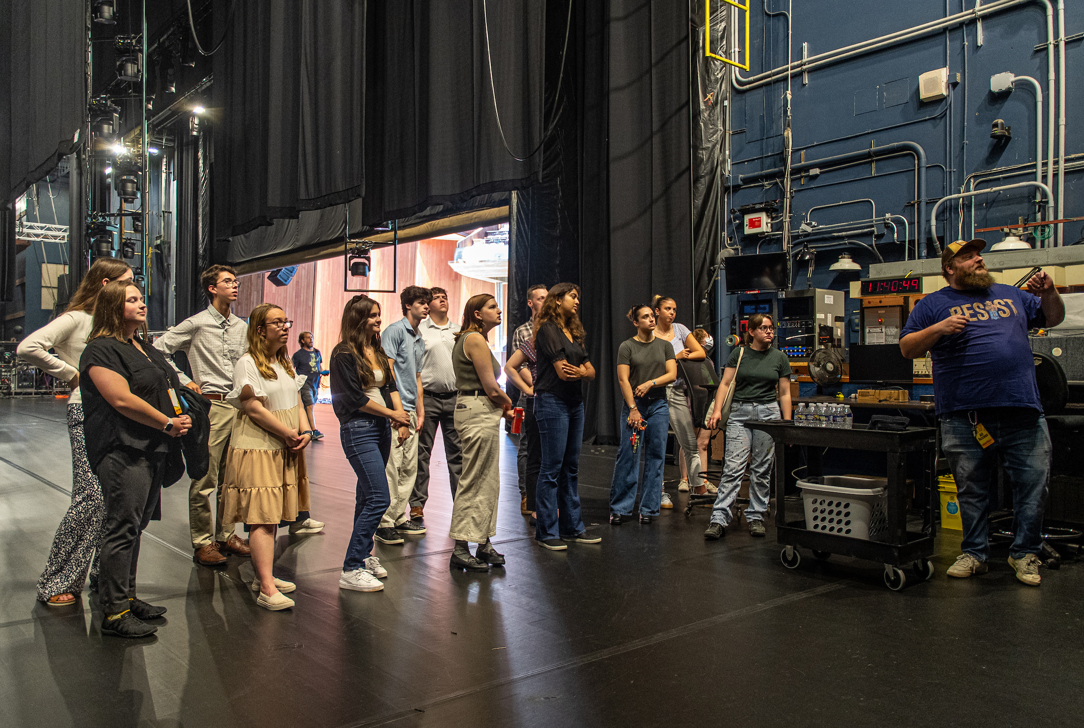 A group of interns stands on the Filene Center stage, listening to a staff member explain backstage equipment and operations. Large curtains and technical gear are visible in the background.