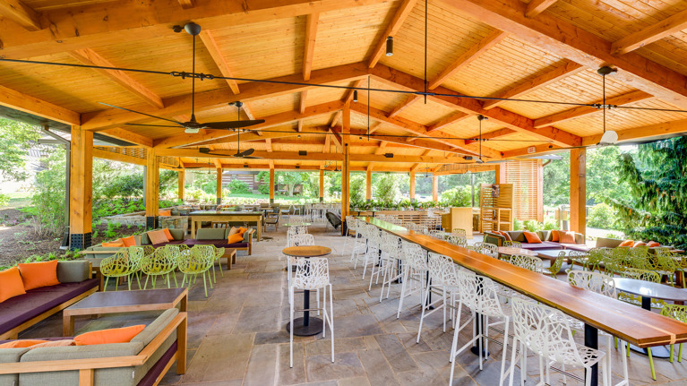 Spacious covered pavilion with wooden beams, ceiling fans, and seating areas featuring tables, chairs, and cushioned benches.