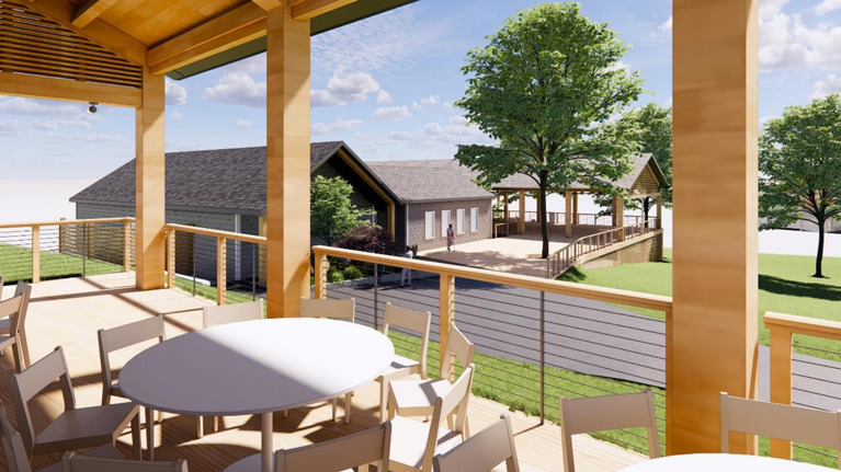 View from inside a wooden pavilion with round tables and chairs, looking out toward a gray building and landscaped area with trees.