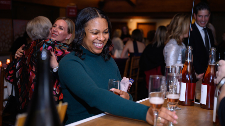 Person in a green outfit smiling and reaching for a glass of sparkling wine at a bar with bottles displayed.