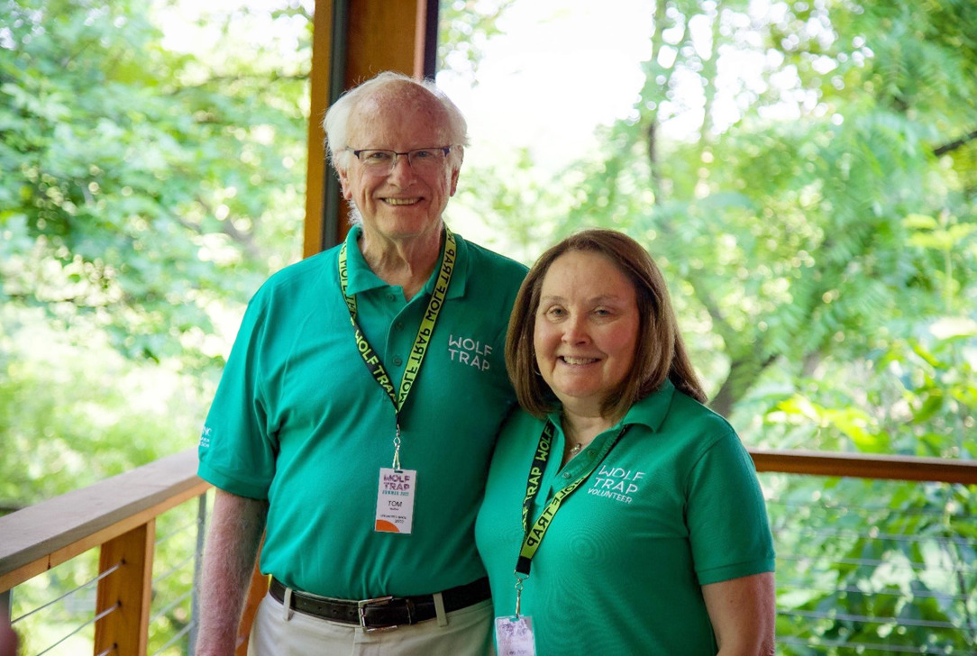 Two Wolf Trap volunteers in green shirts pose on a wood pavilion surrounded by greenery.
