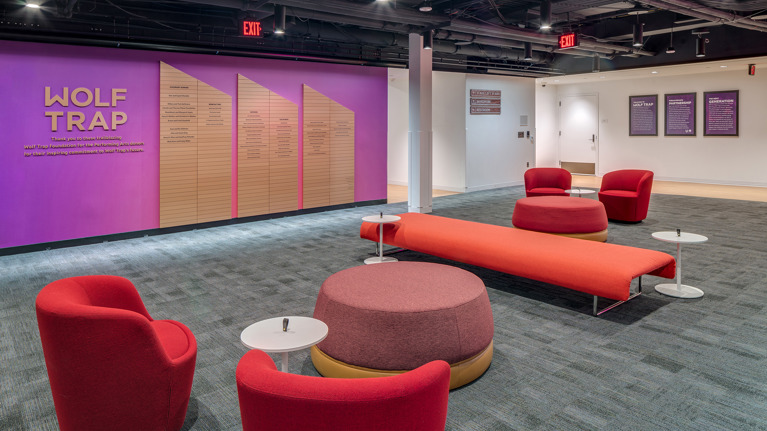 Open seating area with red and pink chairs, circular ottomans, and a purple donor wall in the background.