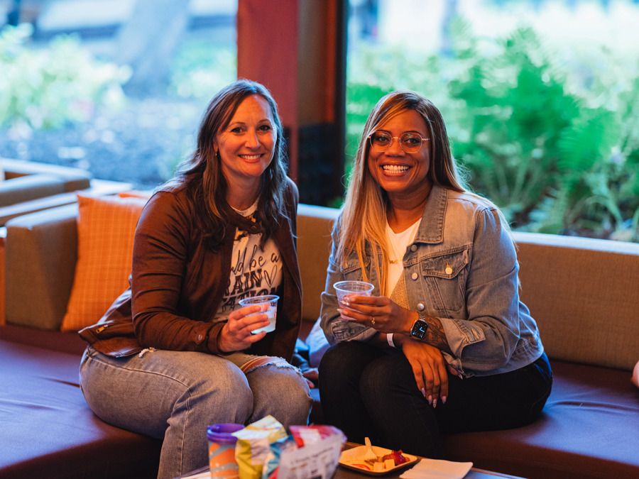 Two friends smile at the camera at Encore Circle Lounge, on a comfy sofa with greenery in the background.