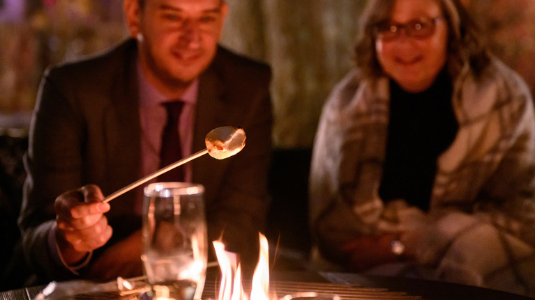 Two people roasting marshmallows by a fire pit, one holding a drink.