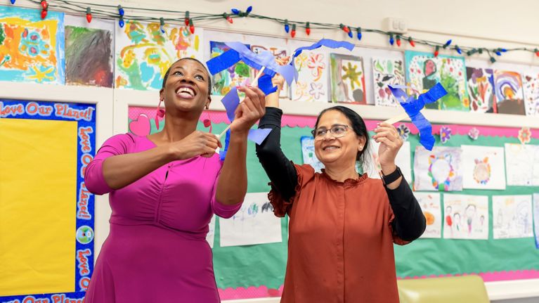 A Wolf Trap teaching artist and classroom teacher standing in a colorful classroom, holding and displaying blue paper craft pieces. The background shows children’s artwork and a bulletin board with bright decorations.