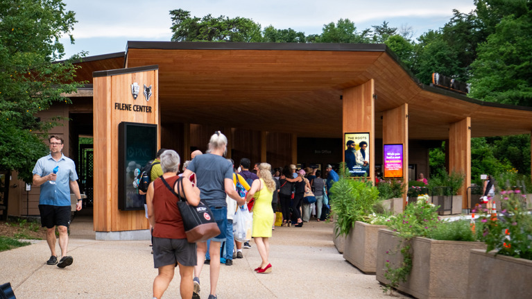 Guests walking toward Filene Center entrance with wood canopy and digital event displays.