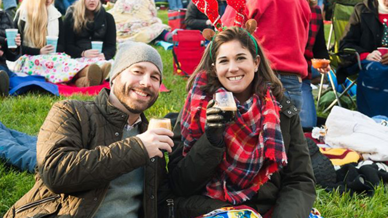 People sitting outdoors on a grassy area during a festive gathering, wrapped in colorful blankets, holding drinks, and surrounded by others in warm clothing and holiday accessories.