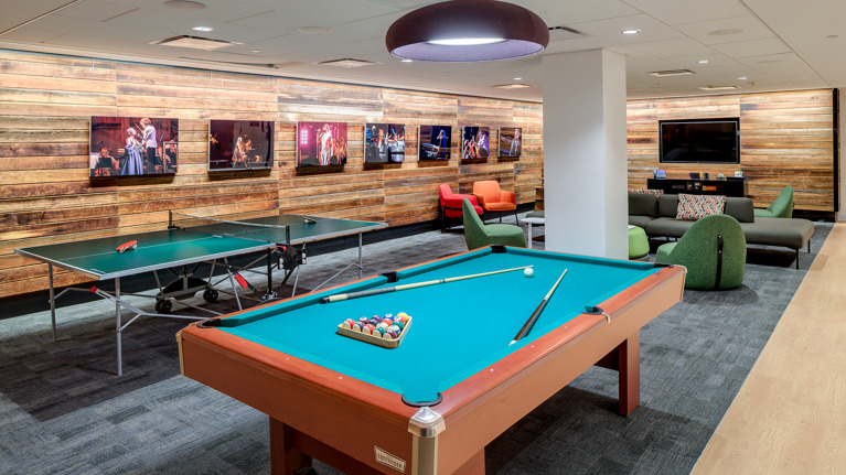Recreation room with pool table, ping pong table, lounge seating, and wood-paneled walls displaying performance photos.