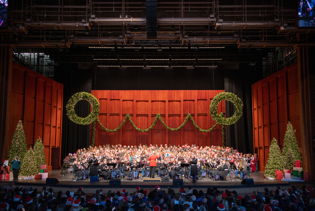 Stage decorated with wreaths and Christmas trees featuring a large choir and musicians performing.