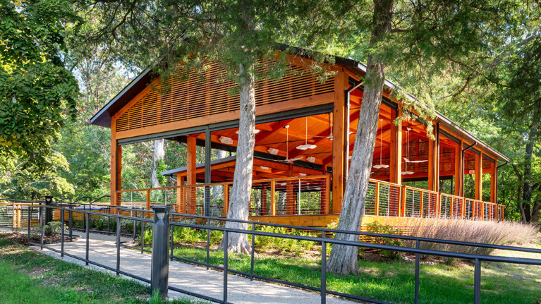 Open-air wooden pavilion surrounded by trees with a sloped roof and railing along a paved path.