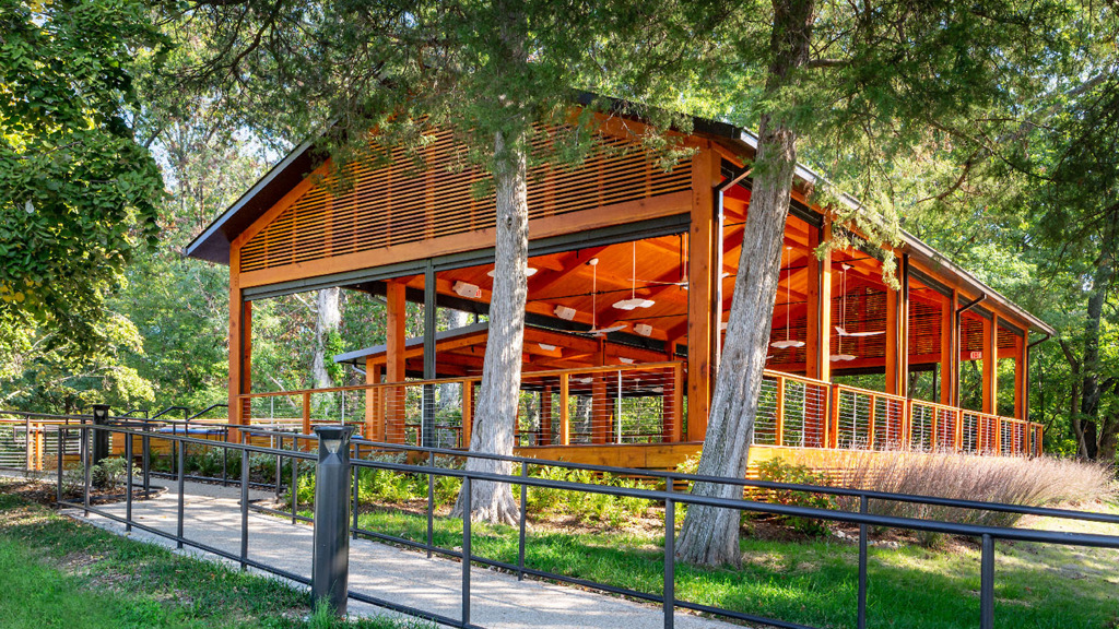 Open-air wooden pavilion surrounded by trees with a sloped roof and railing along a paved path.
