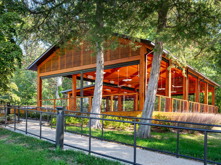 Open-air wooden pavilion surrounded by trees with a sloped roof and railing along a paved path.