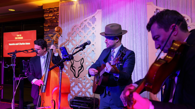 Live band performing with upright bass, guitar, and violin on a stage with event signage.