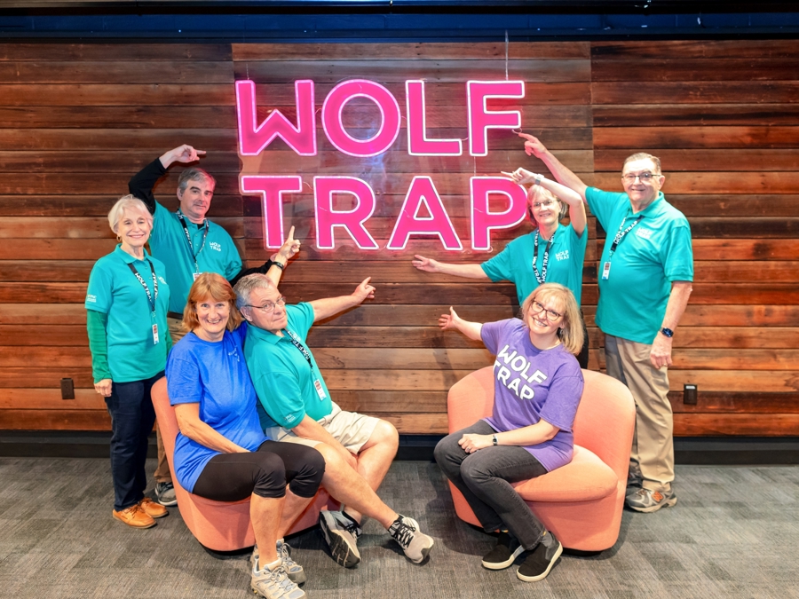 Six volunteers and a Wolf Trap staff member pose in front of a wooden wall with neon "WOLF TRAP" signage, wearing various "WOLF TRAP" shirts.