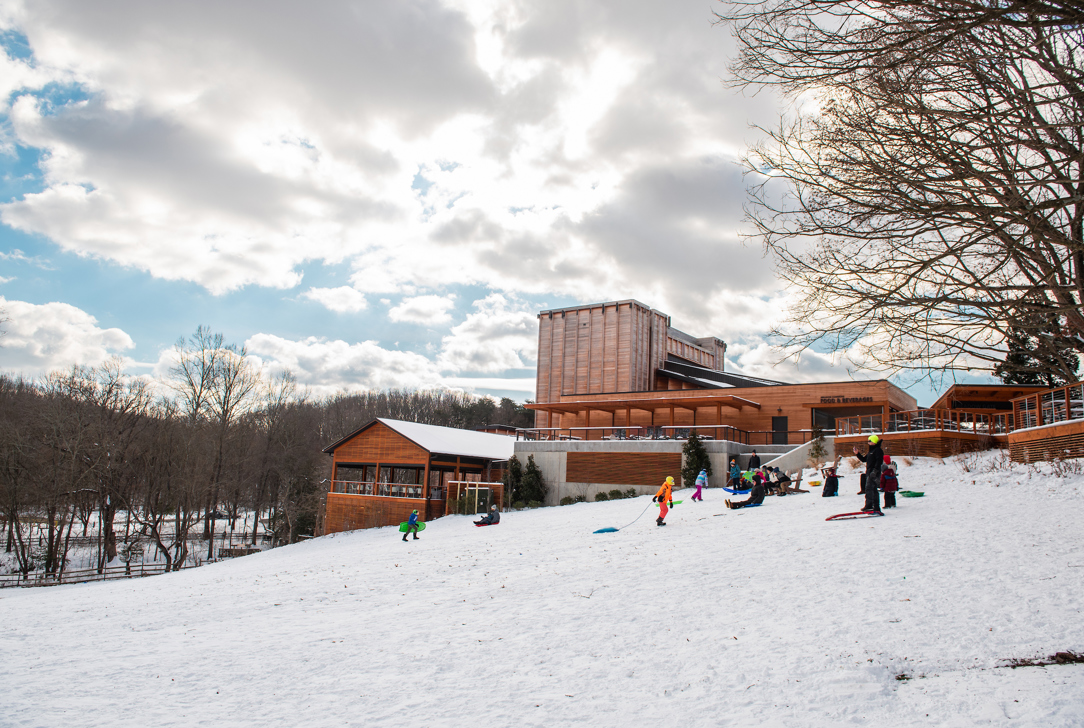 Snow-covered hill with children sledding near the Filene Center under a partly cloudy sky.