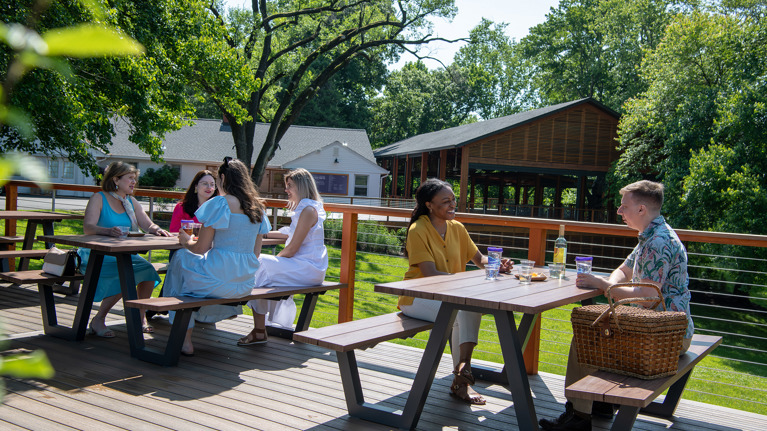 Picnic area on a wooden deck with railing, overlooking green lawn and pavilion in the background.