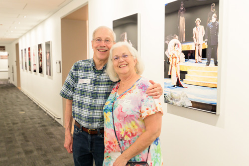 Two people standing in a hallway lined with framed performance photos on the wall.