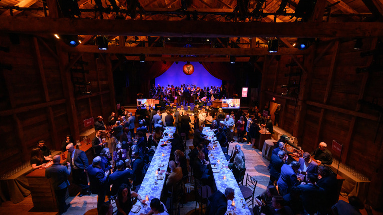 Wide view of a rustic venue with long banquet tables and guests mingling under warm lighting.