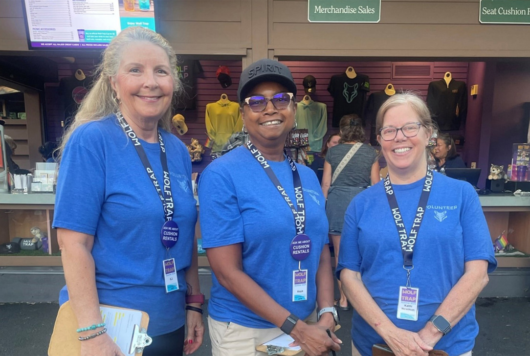 Three volunteers in blue shirts pose in front of the gift shop.
