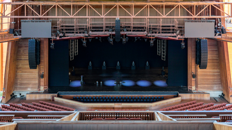 View of Filene Center stage with new large speaker arrays and audio equipment suspended above seating area.