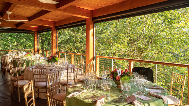 Elegant outdoor dining setup under a wooden pavilion with round tables, green tablecloths, and gold chairs overlooking lush greenery.