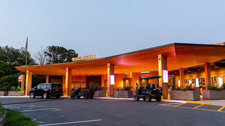 Evening view of Filene Center main gate illuminated with warm lighting and vehicles parked in front.