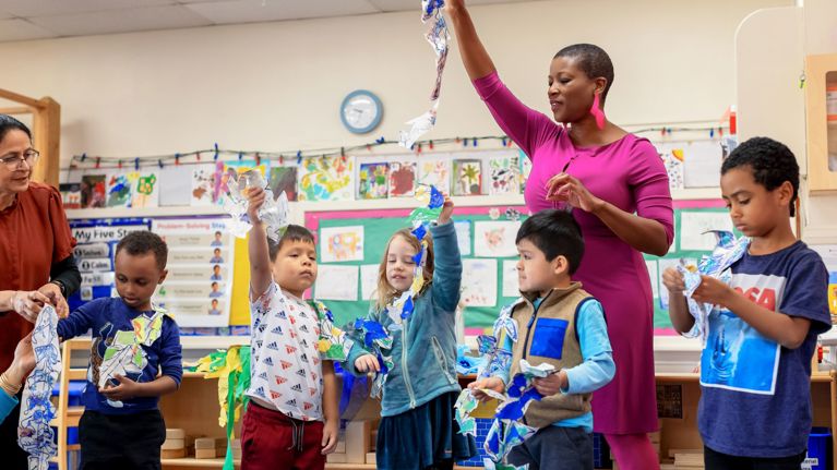 A Wolf Trap teaching artist in a classroom raising a long strip of crumpled paper while several children stand nearby, each holding similar colorful paper pieces. The walls are decorated with children’s drawings and educational posters.