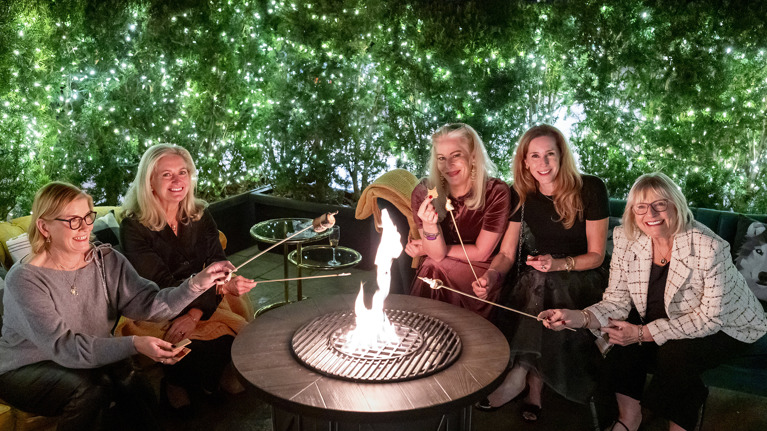 Guests roasting marshmallows over a fire pit surrounded by greenery and string lights.