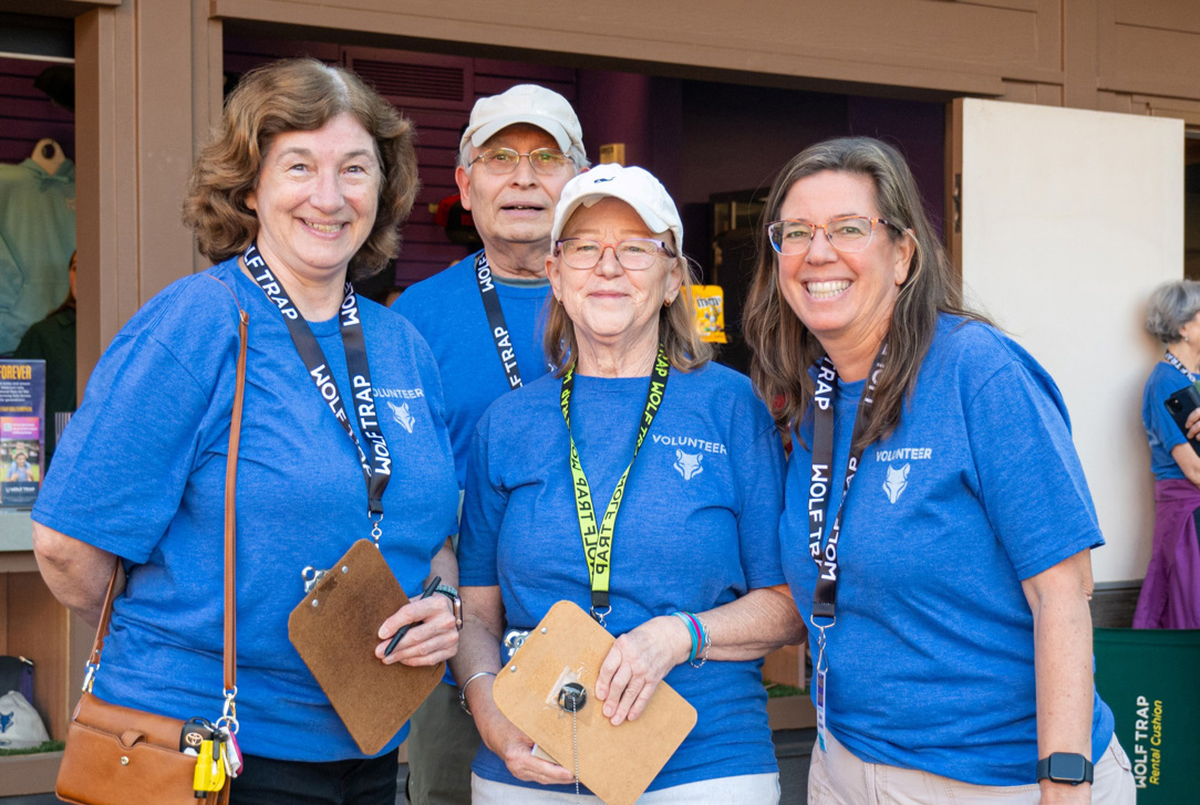 Four volunteers in blue "WOLF TRAP VOLUNTEER" shirts standing outside with clipboards, smiling at the camera.