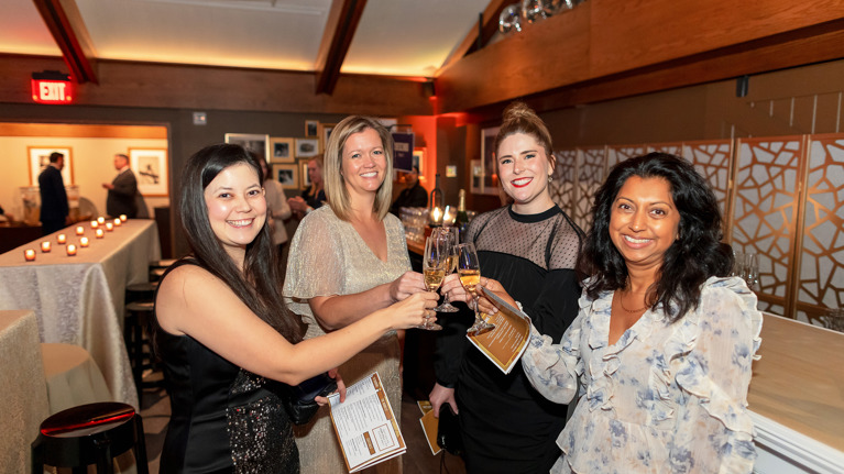 Four women in formal attire toasting with champagne in a candlelit room.