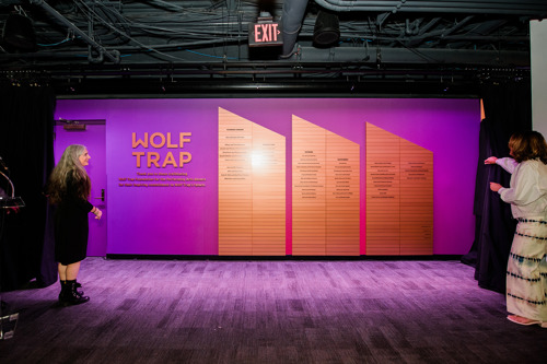 Two people unveiling the Wolf Trap donor wall with purple backdrop and wood panels listing donor names.