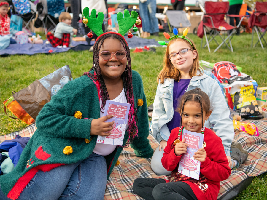 An adult and two children sitting on a picnic blanket outdoors wearing holiday-themed headbands and holding event programs.