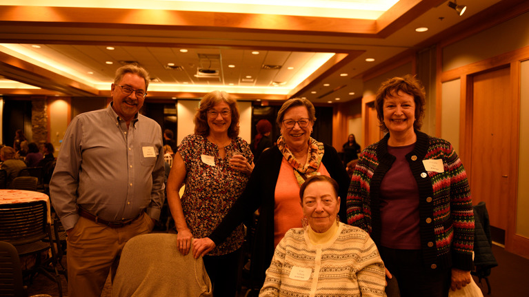 Five volunteers posing together in the event space, with tables and other guests visible in the background.