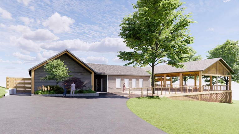 Exterior view of a gray building with pitched roof and landscaped entrance, next to a spacious wooden pavilion with railing and trees in the background.